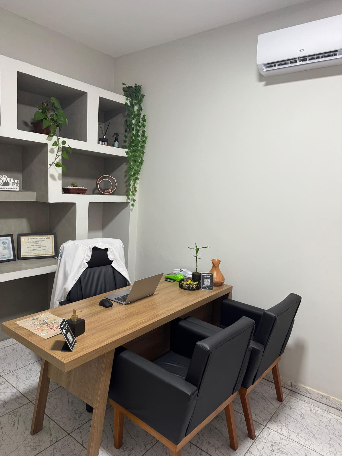 Sala de atendimento - vista da mesa com jaleco, poltronas e armário de nichos com diplomas