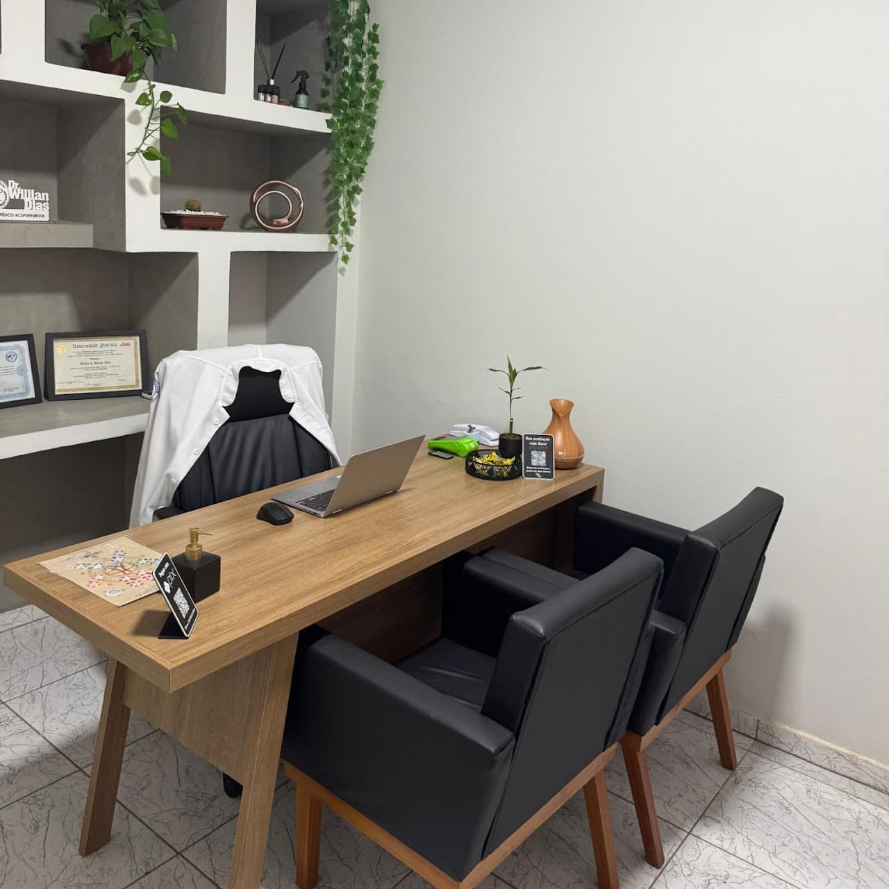 Sala de atendimento - vista da mesa com jaleco, poltronas e armário de nichos com diplomas