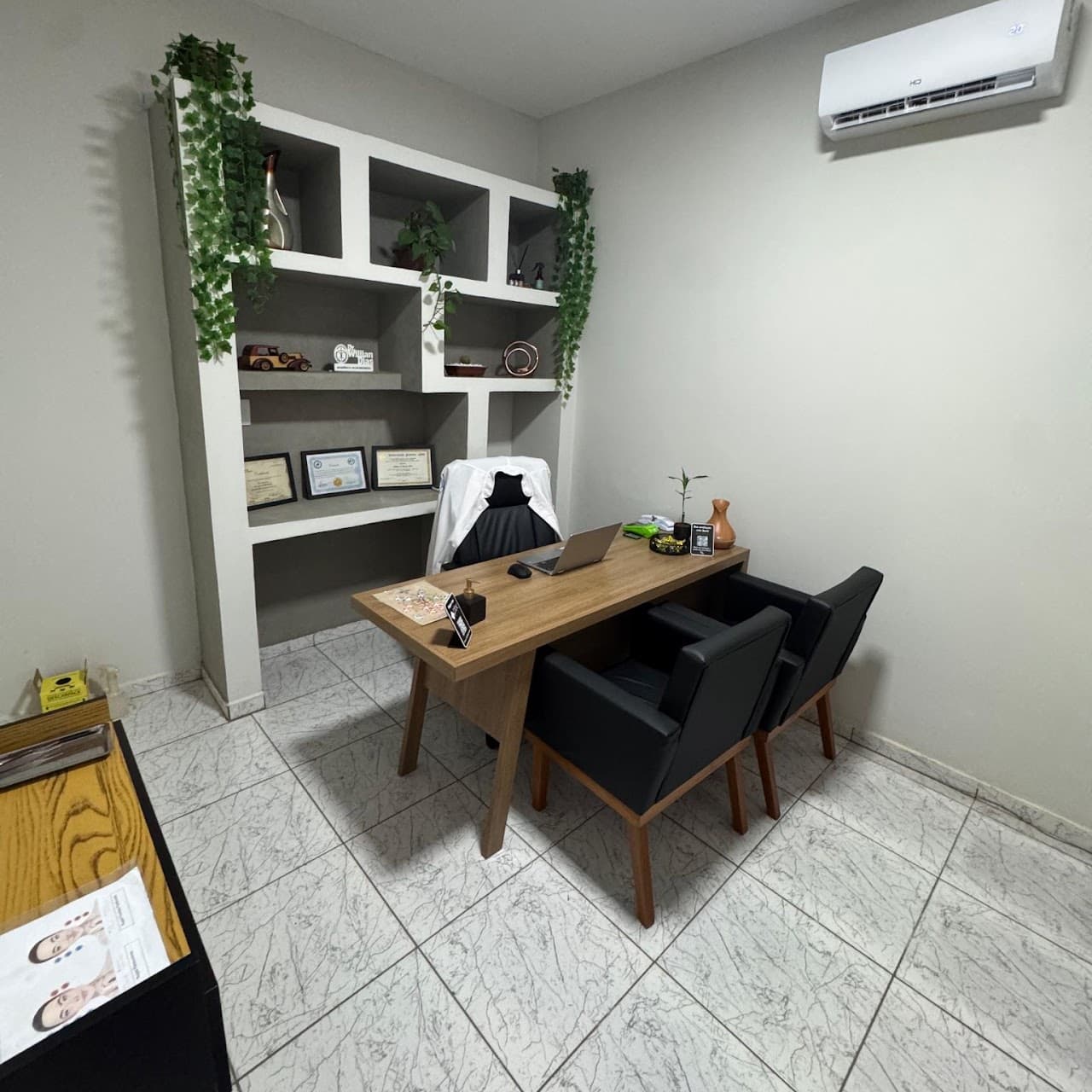 Sala de atendimento - visão ampla com armário de nichos, diplomas e maca ao fundo à esquerda